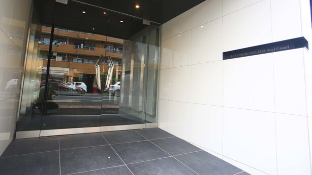 A modern building entrance with a glass door, a sign reading "AXAS ISHIKAWA-DAI Asyl Court," and a reflection of a multi-story building and parked cars. - 2500 Comics Room (Tokyo)
