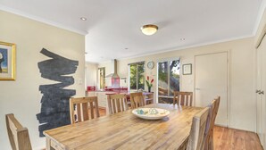 A dining area with a wooden table and chairs, a black abstract painting on the wall, and a kitchen in the background. - All Decked Out (Port Willunga)