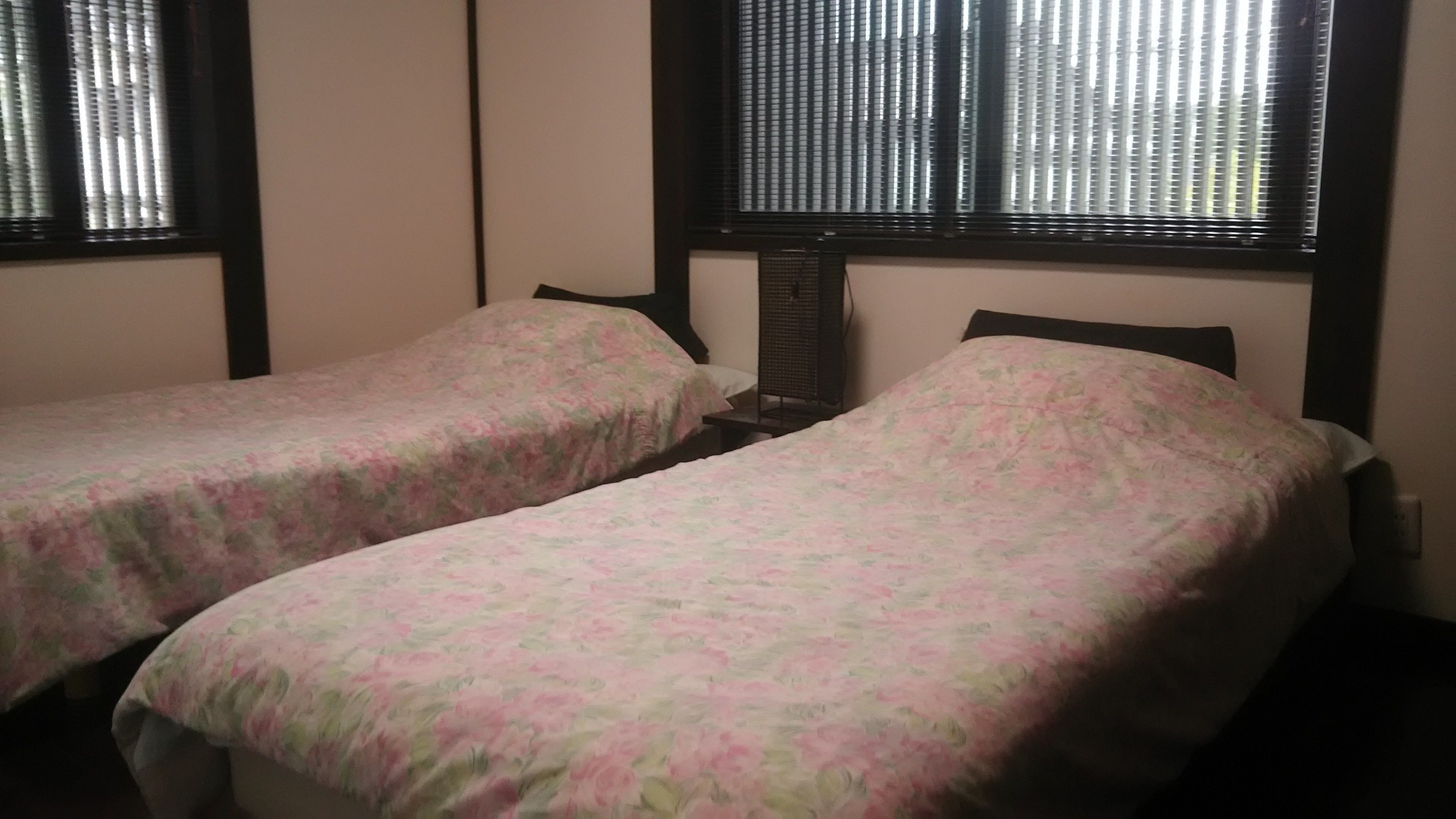 A room with two single beds, each with a floral-patterned bedspread. There is a window with blinds on the wall behind the beds.