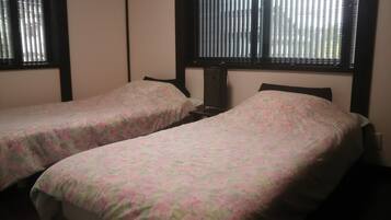 A room with two single beds, each with a floral-patterned bedspread. There is a window with blinds on the wall behind the beds.