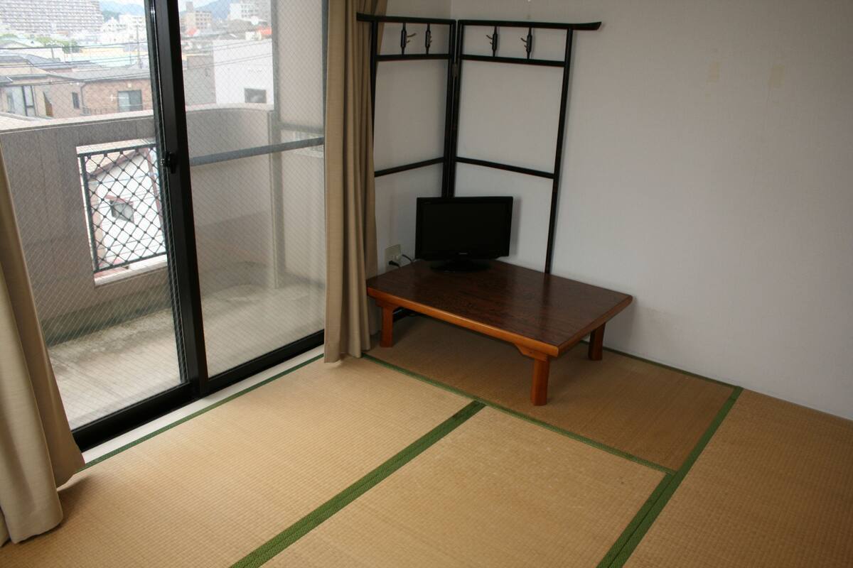 Une pièce avec un sol recouvert de tatamis, une table en bois avec un téléviseur et une porte-fenêtre coulissante donnant sur un balcon.