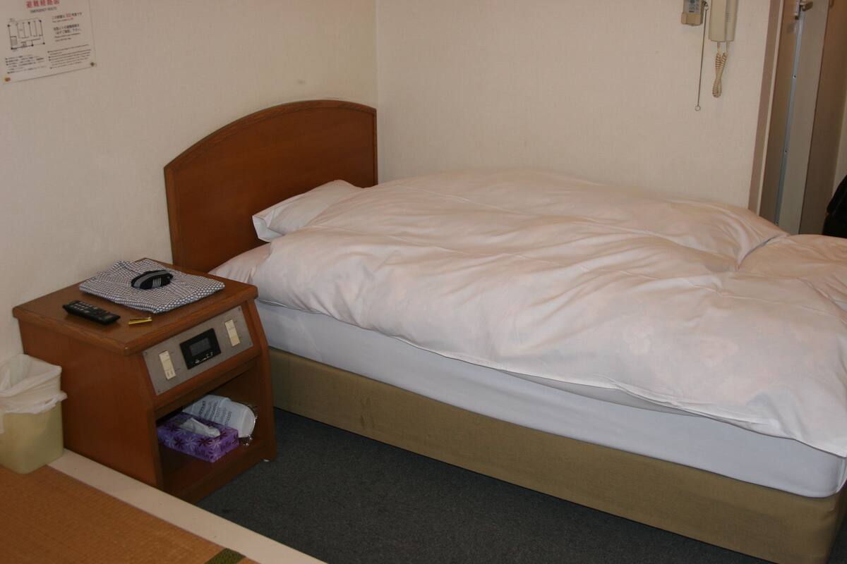 Un lit simple avec une tête de lit en bois, une table de chevet avec une télécommande, une boîte de mouchoirs et une petite étagère sur laquelle se trouvent une boîte de mouchoirs et un objet violet.