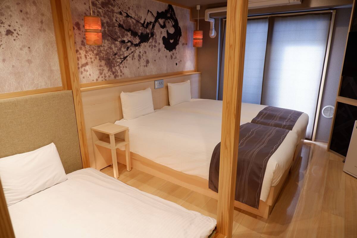 A hotel room with two beds, a small bedside table, a wall-mounted light, and a window with blinds.