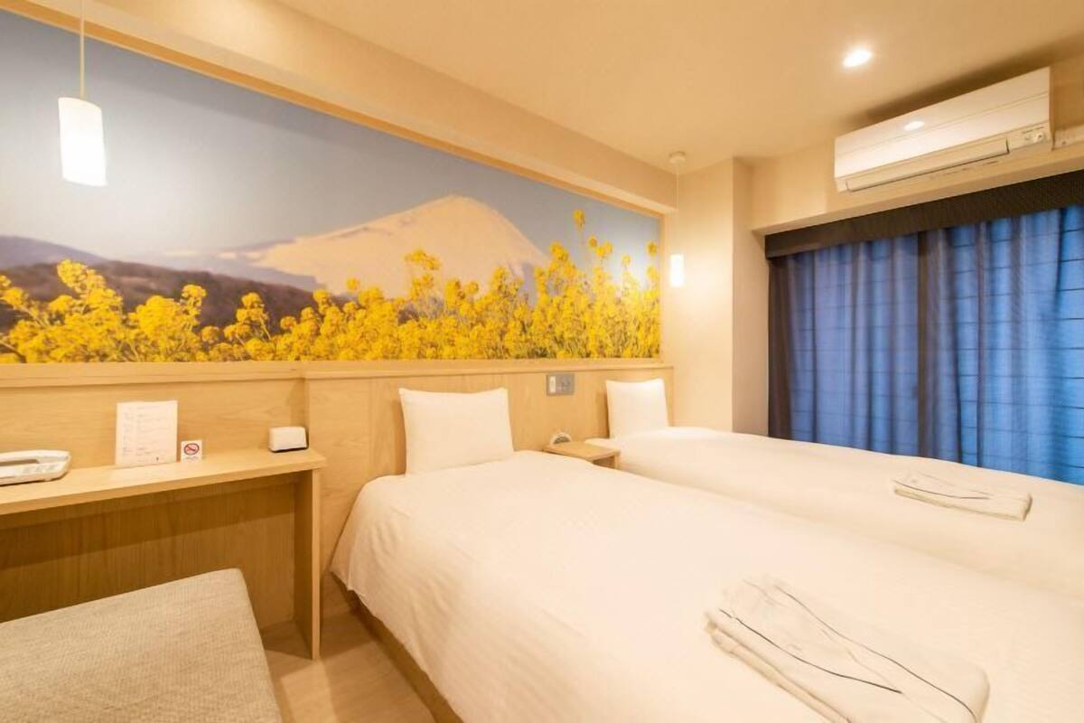 A hotel room with two beds, a desk with a telephone, a wall-mounted air conditioner, and a large window with blue curtains.