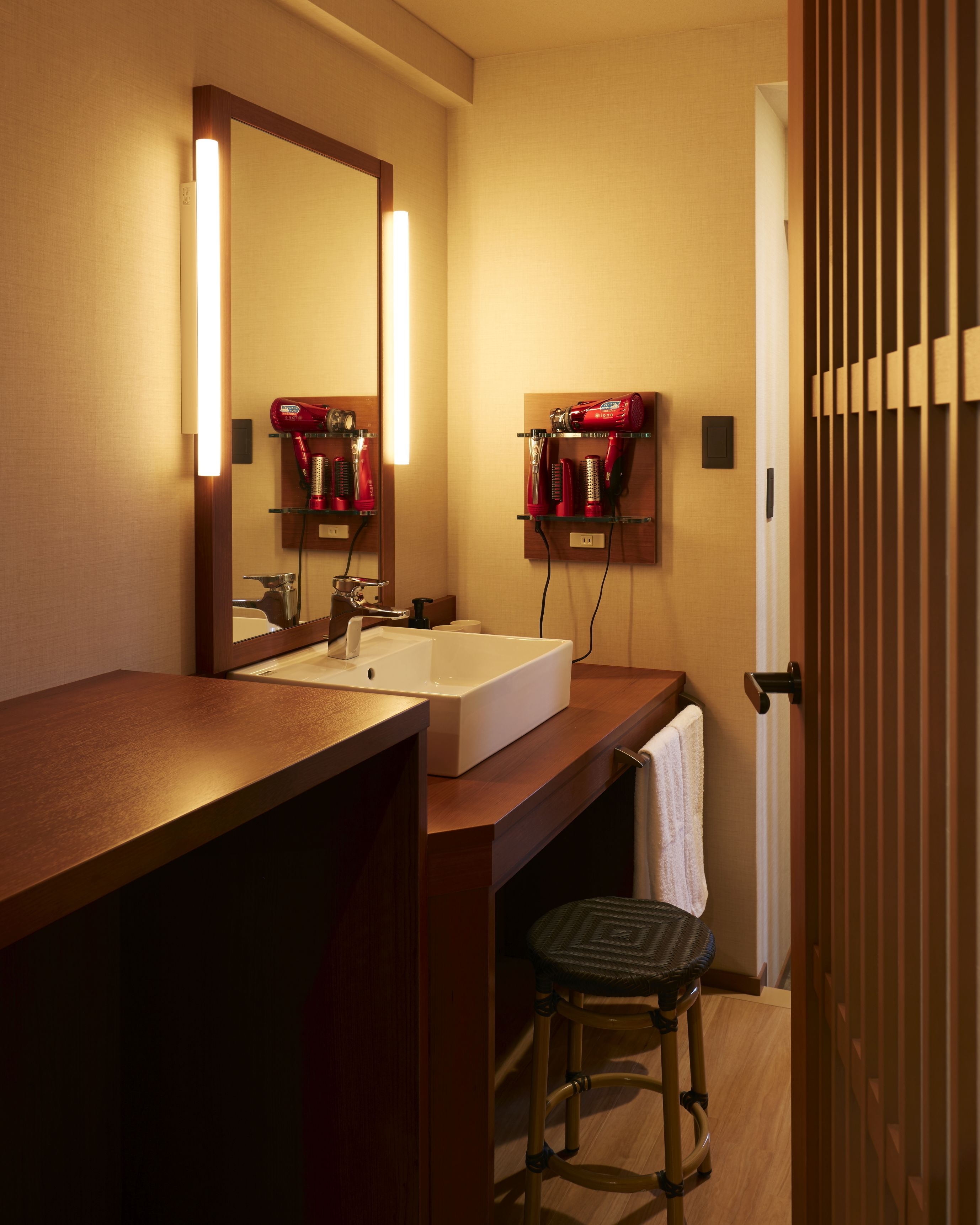 Un baño con tocador de madera, un lavabo blanco, un espejo y un secador de pelo rojo sobre una estantería.