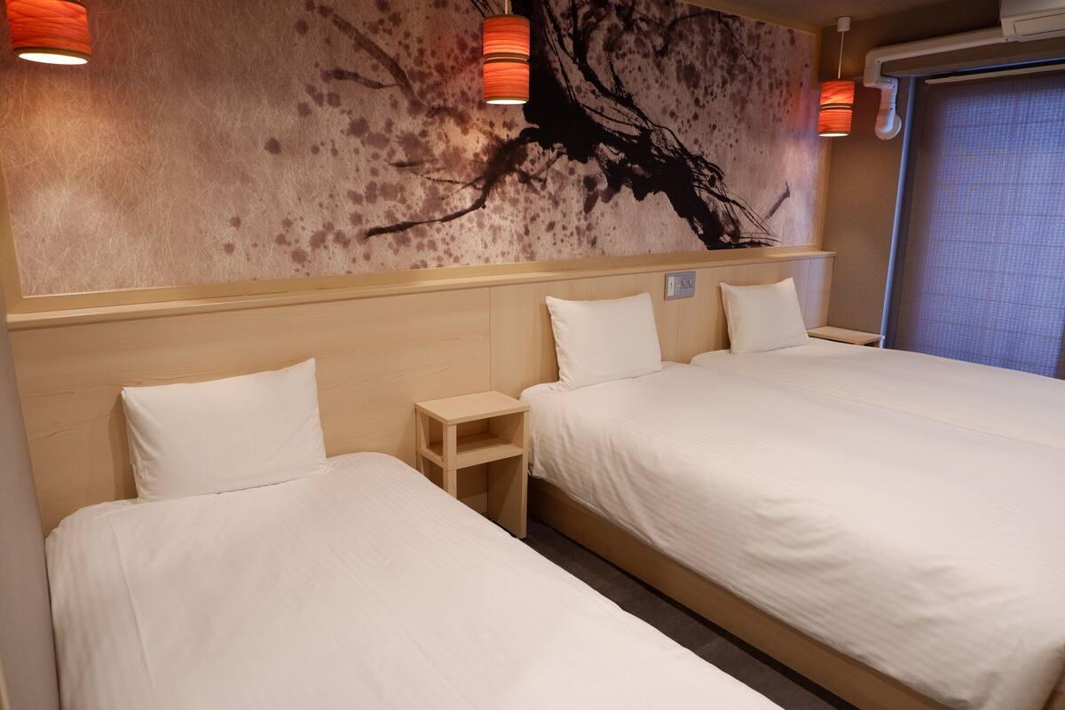 A hotel room with two beds, a wooden nightstand, and a wall with a tree branch mural.