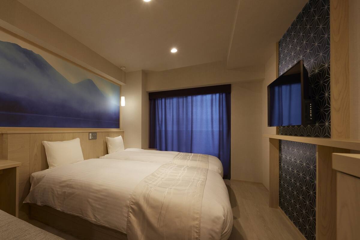 A hotel room with a large bed, a bedside table with a lamp, a painting of a mountain landscape, and a view of the outside through a window with blue curtains.