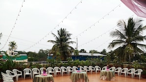Un espace extérieur aménagé pour les événements, avec des tables rondes et des chaises en plastique blanc, entouré de palmiers et illuminé de guirlandes lumineuses.