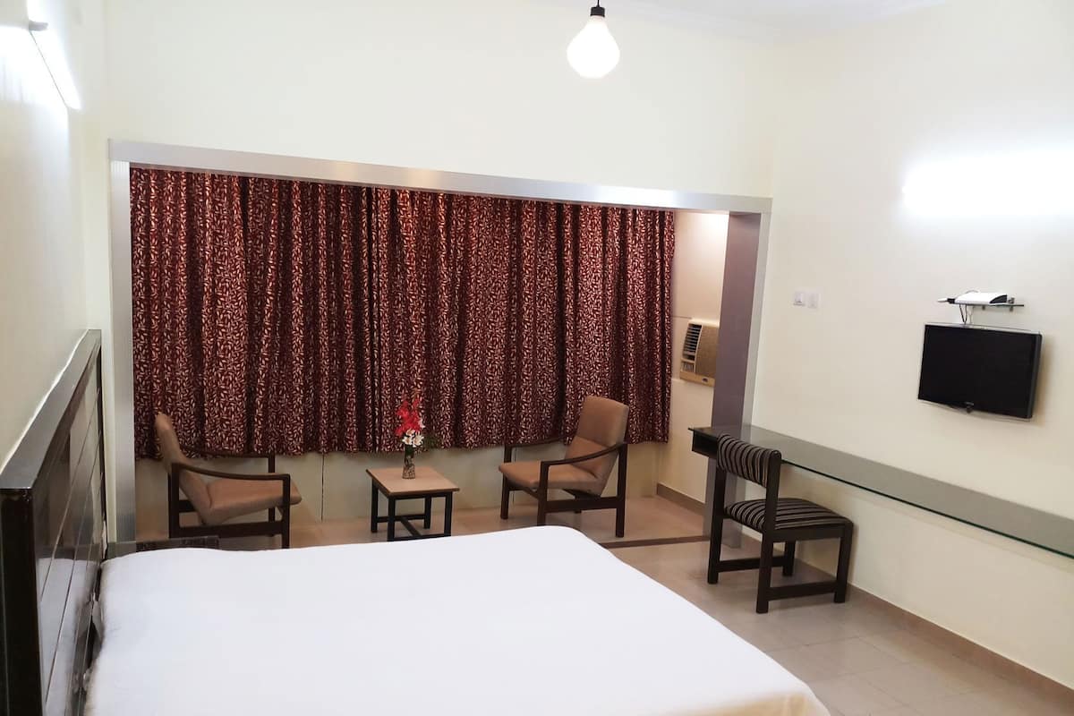Habitación de hotel con cama, dos sillas, una mesita con un florero, televisor y ventana con cortinas estampadas.