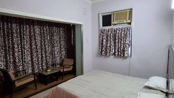A bedroom with a bed, a chair, a table, a ceiling fan, and a window with curtains.