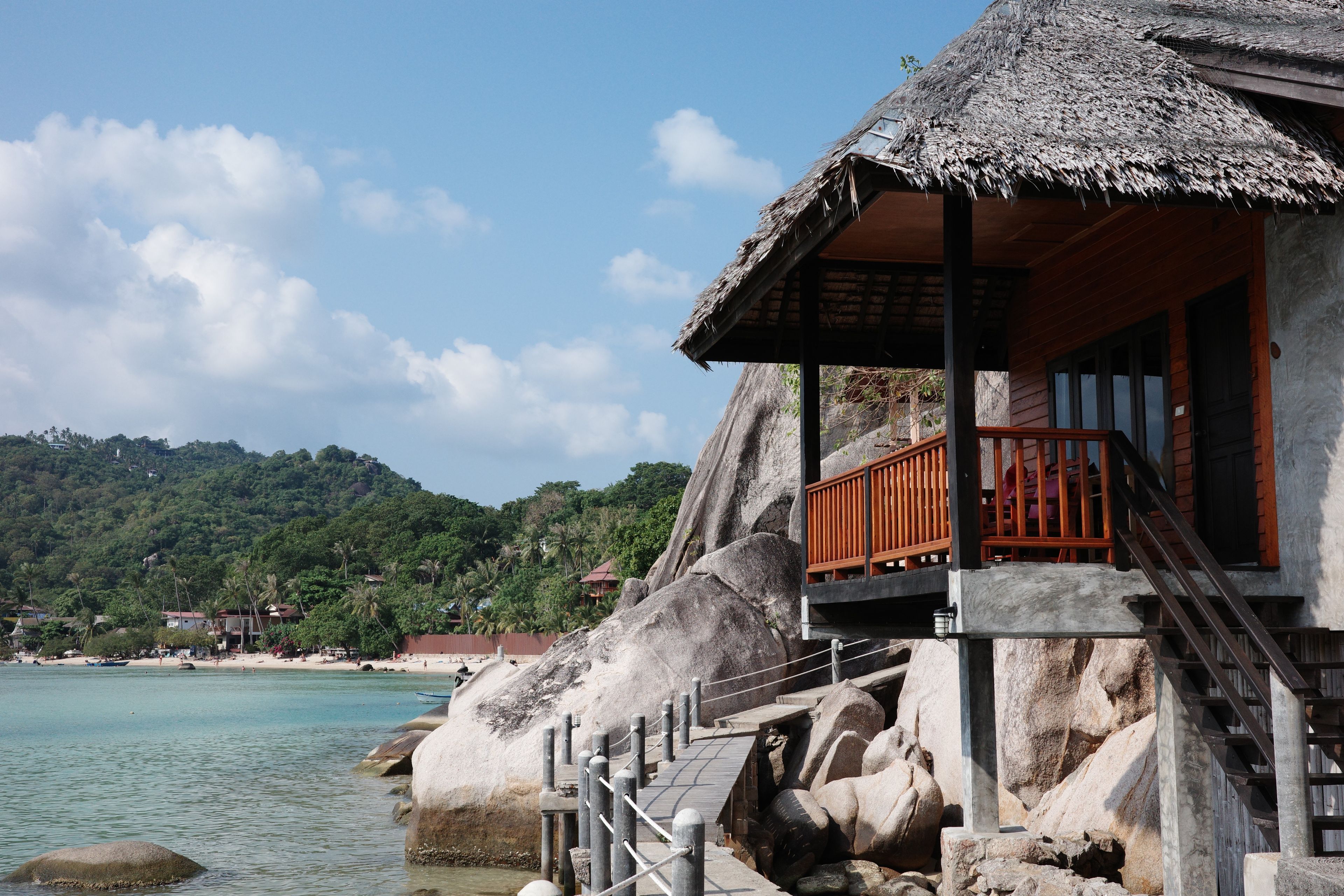 Une propriété située en bord de mer, dotée d’une structure à toit de chaume, d’une terrasse en bois et offrant une vue sur la mer et les collines.