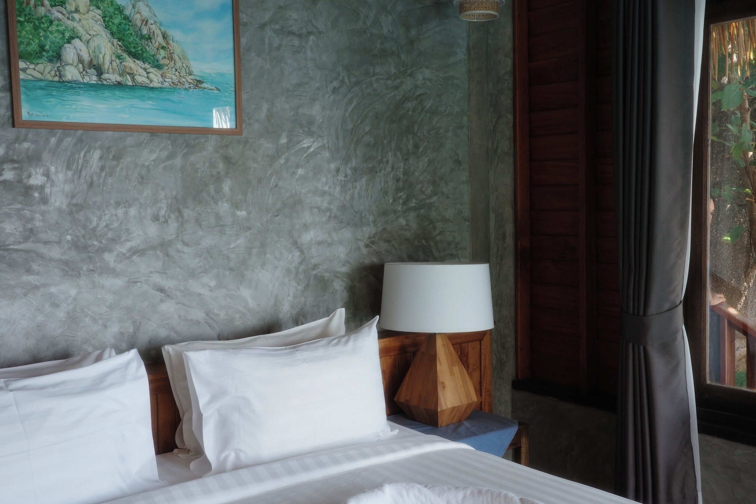 A bed with white linens, a wooden nightstand with a lamp, a painting of a coastal scene, and a window with curtains.