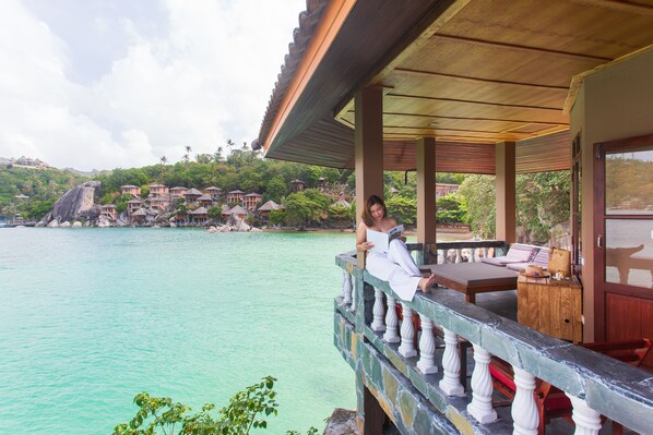 A balcony with a view of turquoise water, a wooden deck, and a building with a traditional roof.
