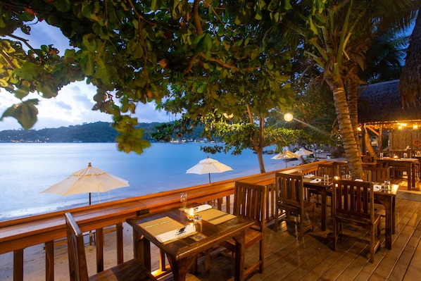 An outdoor dining area with wooden tables and chairs, umbrellas, and a view of the sea.
