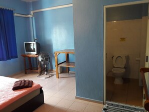 A hotel room with a bed, a TV, a fan, a small table, and a bathroom visible through an open door. - JP Hometel (Krabi)