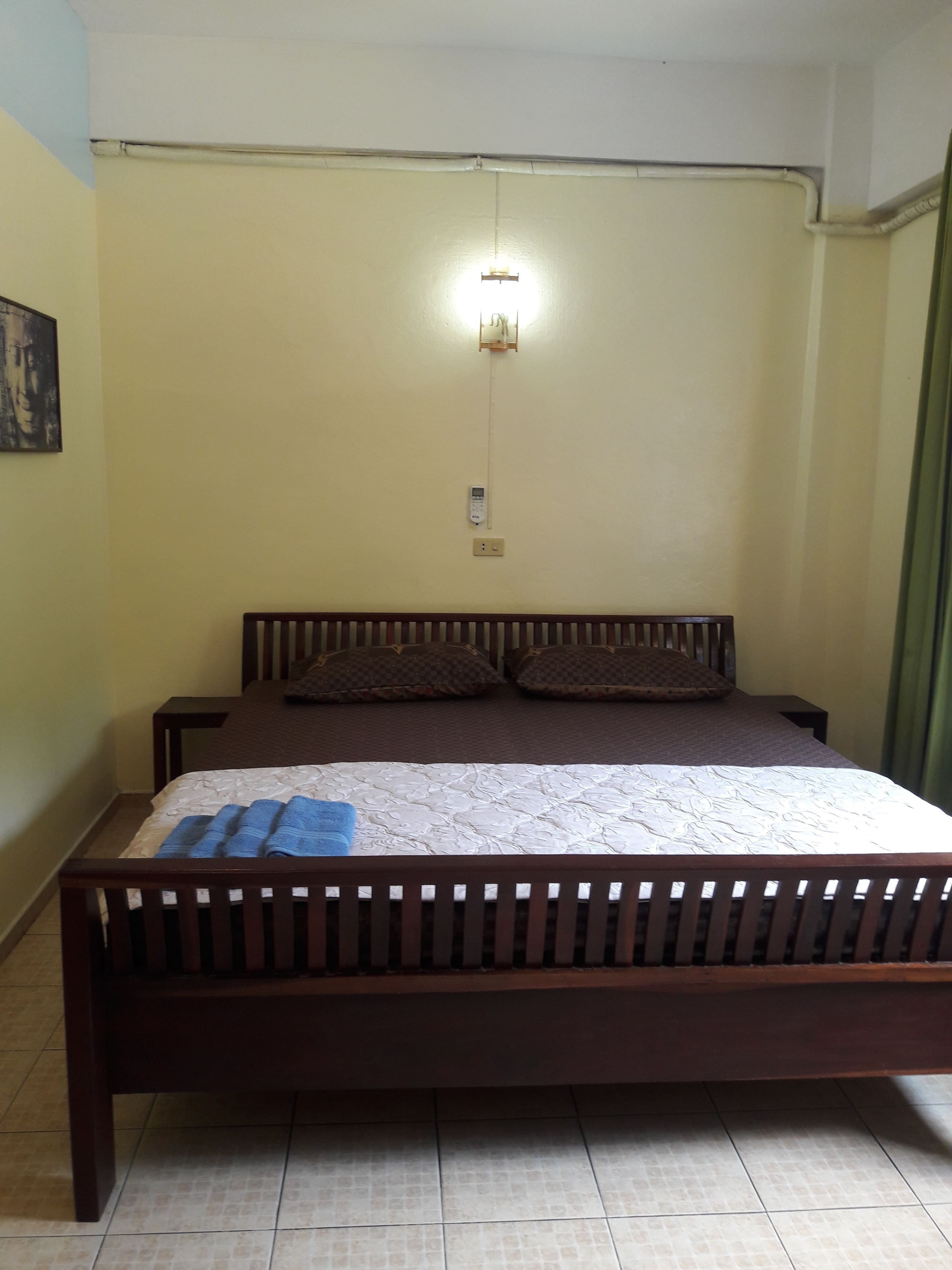 A bedroom with a wooden bed, a nightstand, a wall-mounted light, and a power outlet.
