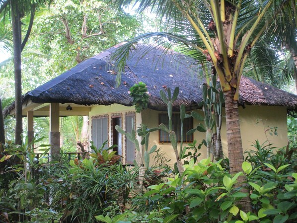 A thatched-roof structure with a covered porch, surrounded by lush tropical vegetation.