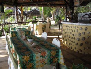An outdoor dining area with tables covered in floral tablecloths, a thatched roof, and a wall decorated with shells.