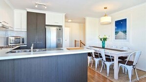 A modern kitchen with white cabinetry, a central island, and a dining area with a table and chairs. - Sunset at Snapper Point (Aldinga Beach)