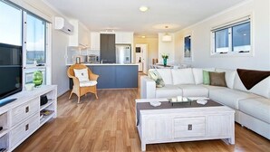 A modern living room with a sofa, coffee table, and TV stand. There is a kitchen area in the background with a dining table and chairs. - Sunset at Snapper Point (Aldinga Beach)