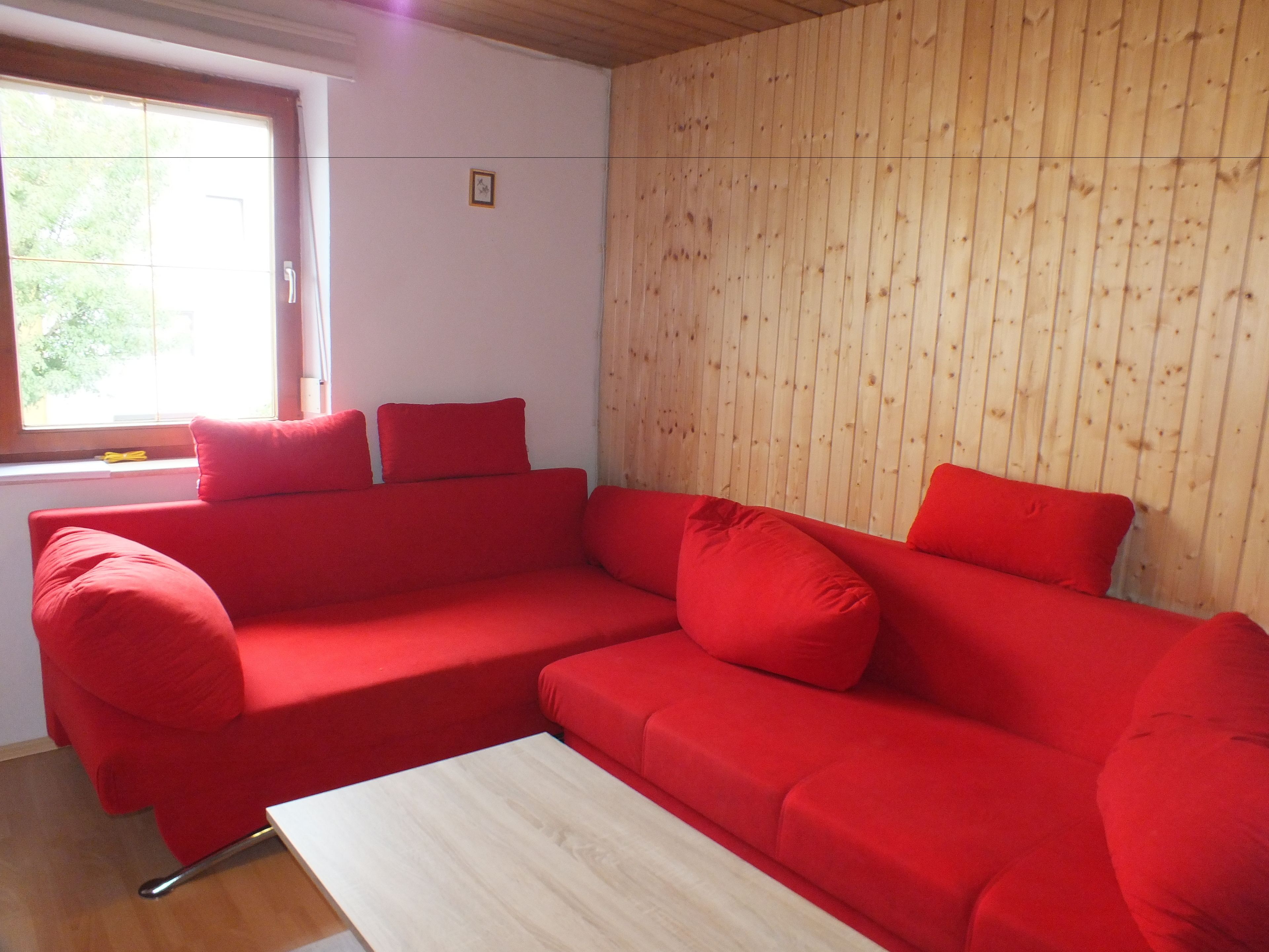 A red sectional sofa with a wooden floor and a window with curtains.
