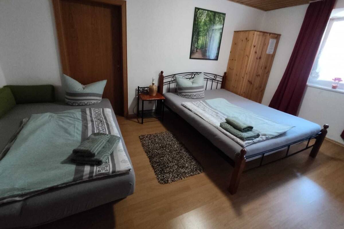 Ein Zimmer mit zwei Einzelbetten, einem Holzschrank, einem kleinen Tisch mit Lampe sowie einem Fenster mit Vorhängen.