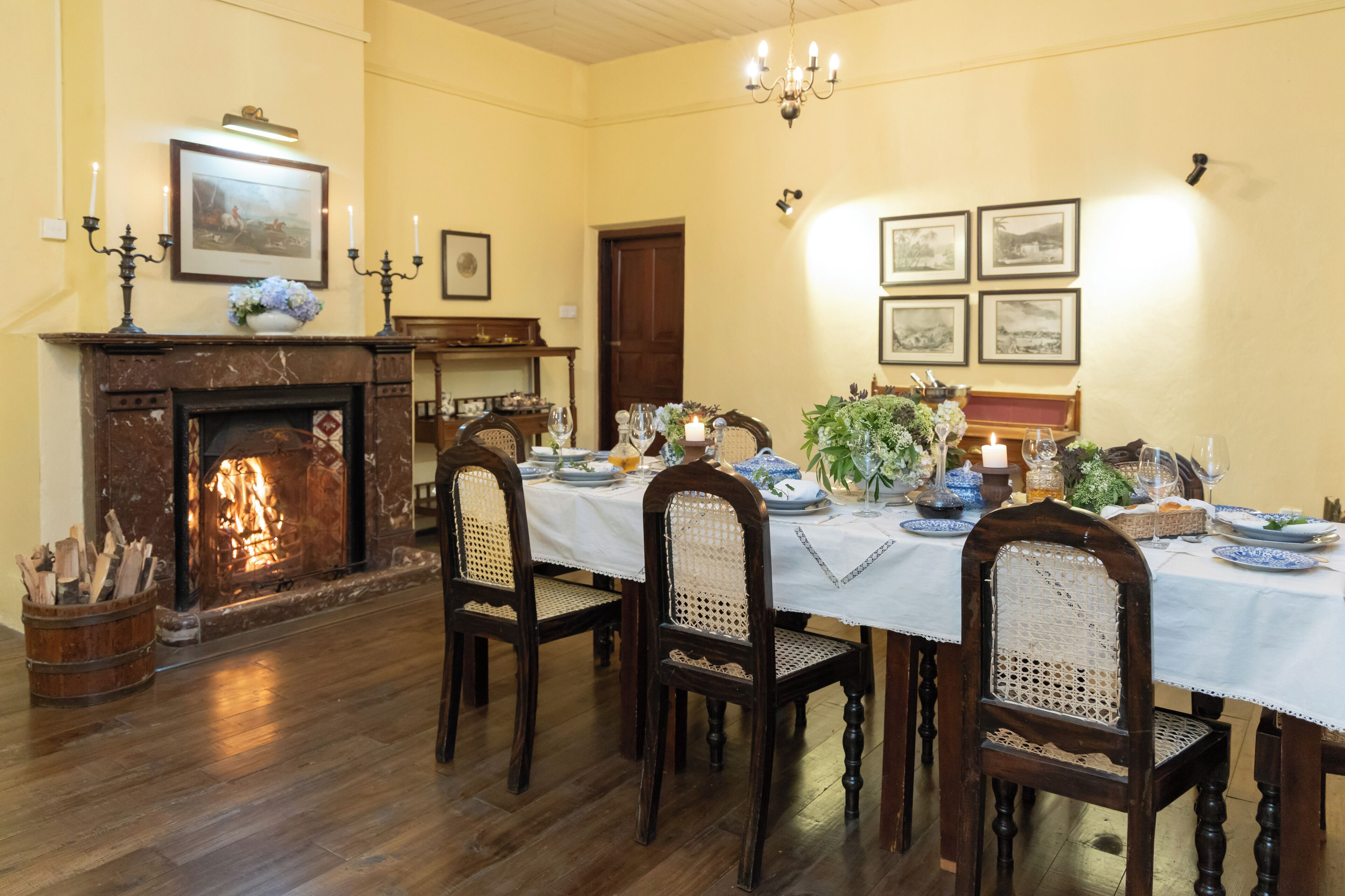 A dining room with a fireplace, wooden chairs, a table set for a meal, and framed pictures on the wall.