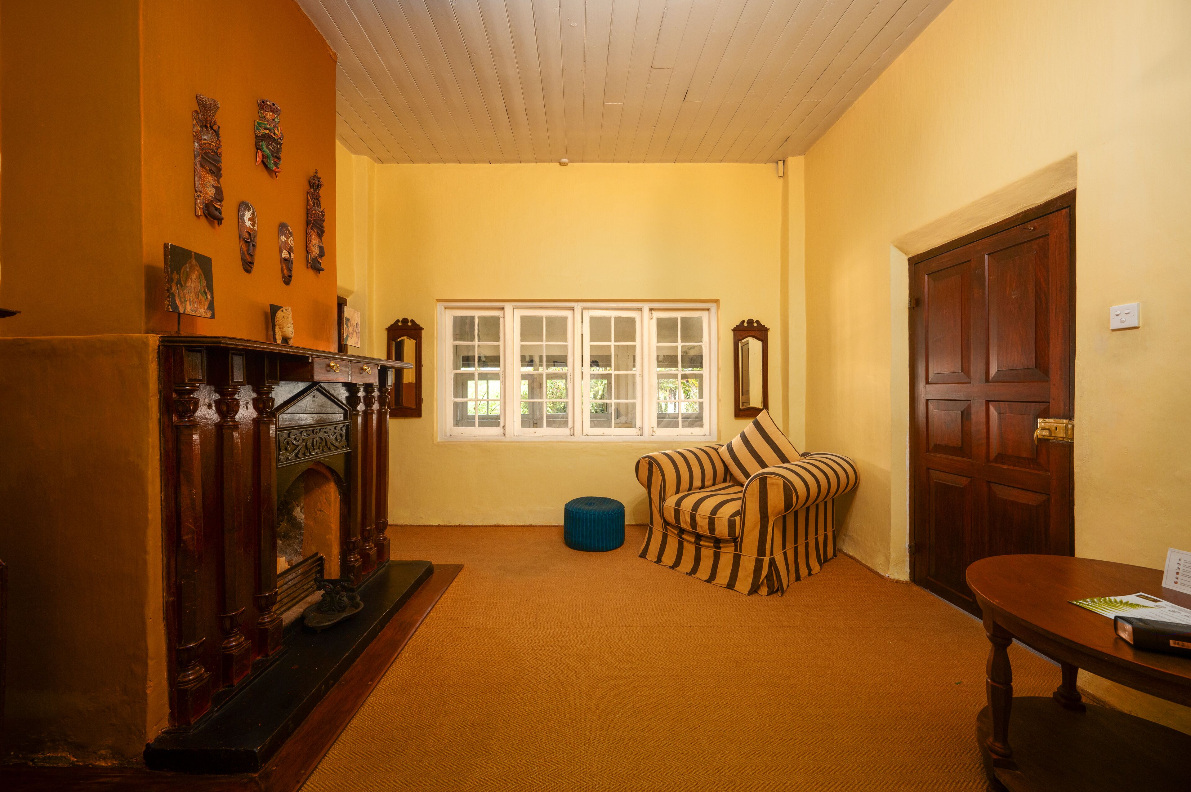 Una habitación con chimenea, un sillón a rayas, una mesa de madera y un ventanal con marcos blancos.