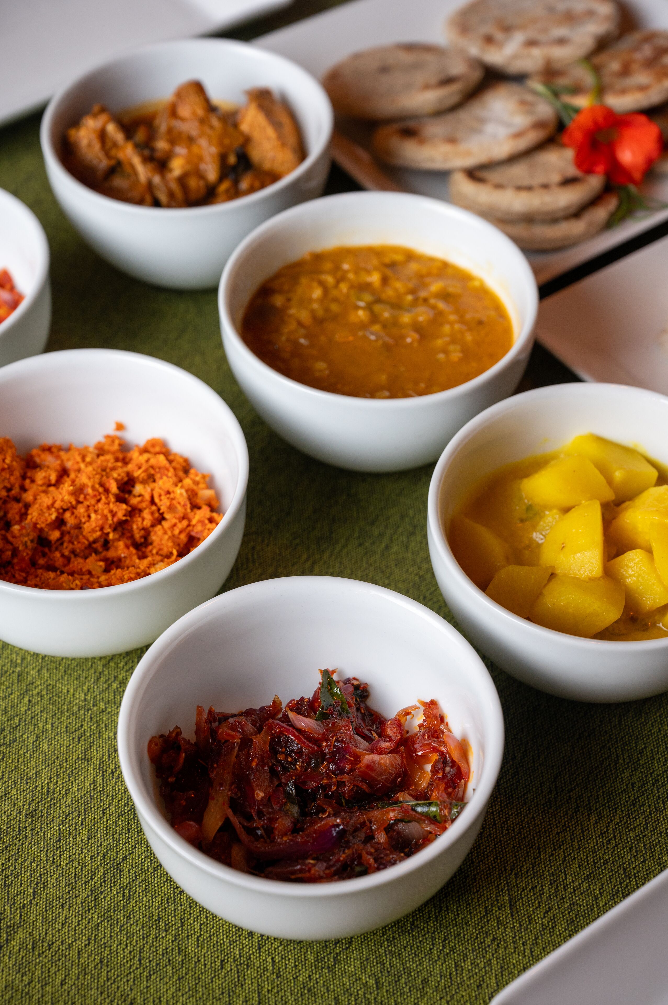 On peut voir une variété de plats servis dans des bols, dont un plat qui ressemble à un curry, un plat à base de mangue et un plat rouge et épicé, le tout disposé sur un tissu vert.