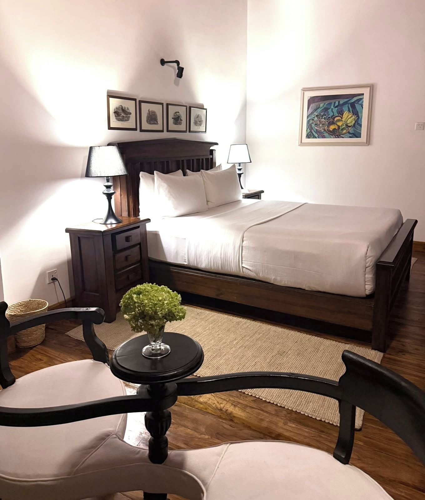 Un dormitorio con cama, mesitas de noche, una lámpara, una silla, una pequeña mesa con una planta y obras de arte en las paredes.
