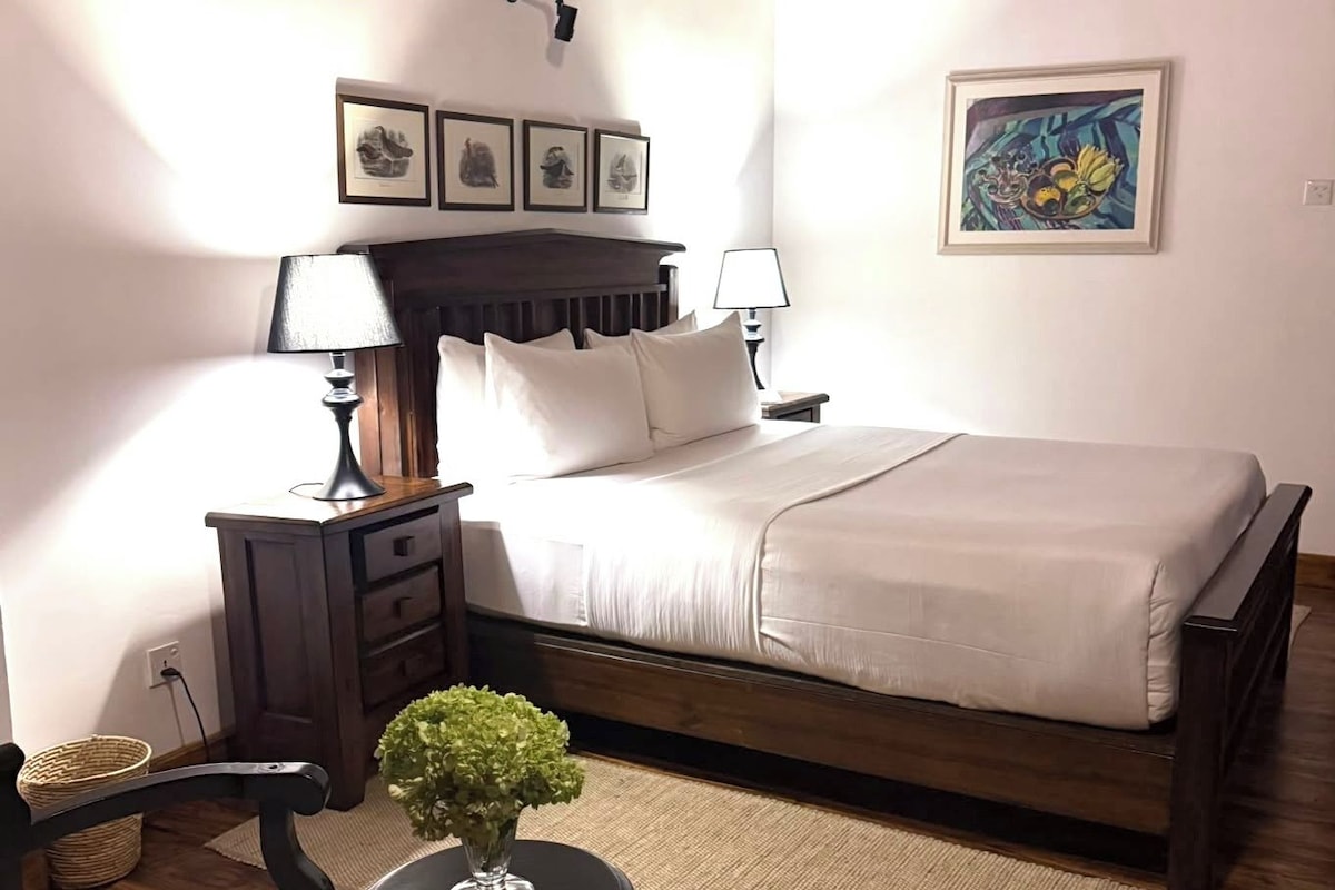 A bedroom with a bed, bedside tables, a lamp, a chair, a small table with a plant, and artwork on the walls.
