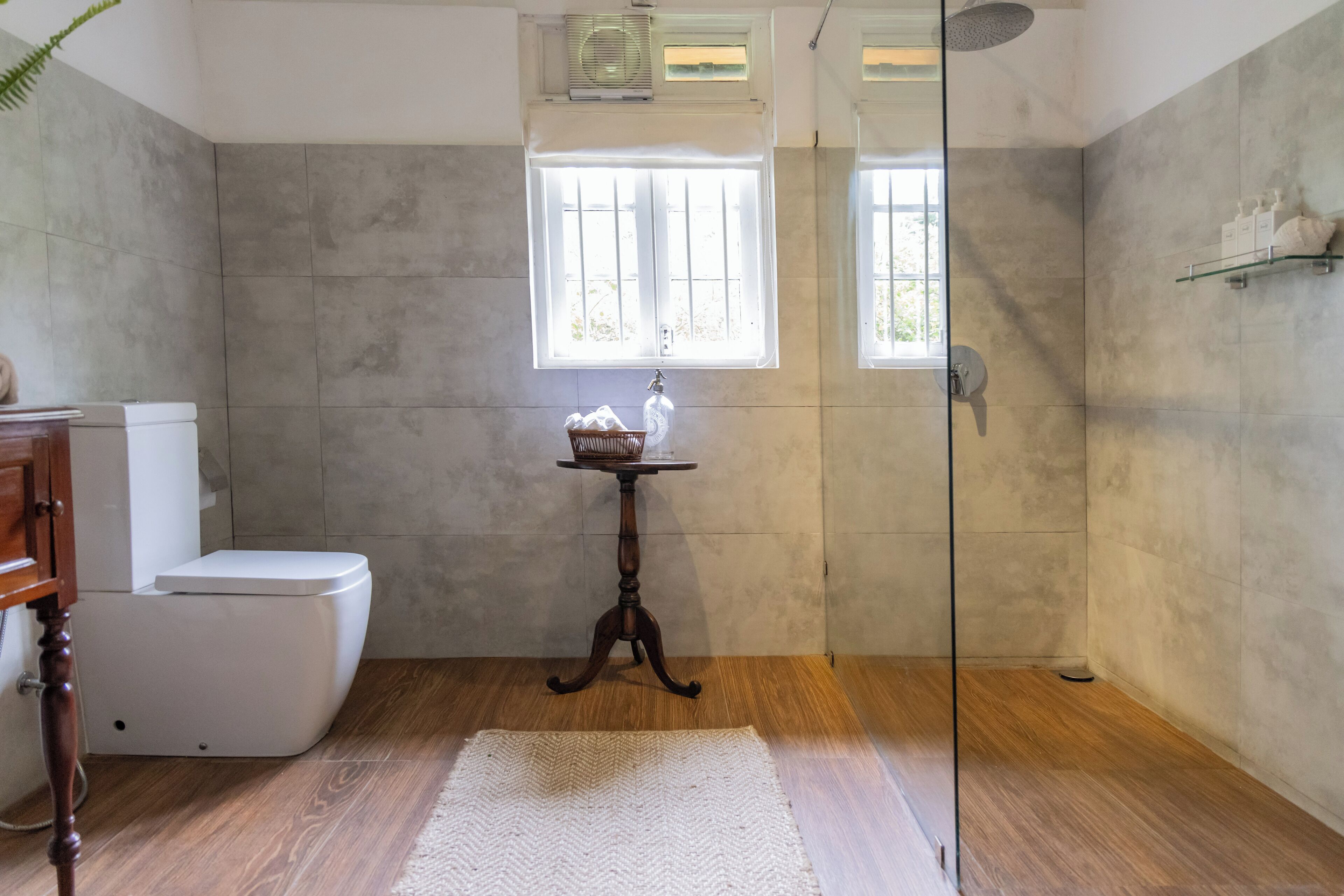 A bathroom with a toilet, a small table with toiletries, a glass shower enclosure, and a window with a view.