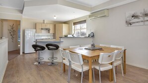 A modern kitchen with white cabinets, a wooden dining table, and white chairs. There is a refrigerator, a sink, and a window with a view of the outdoors. - The Lighthouse (Port Willunga)