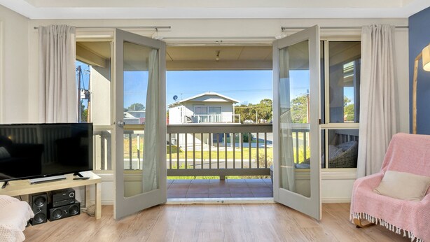 A room with a television, a sofa, and a balcony with a view of houses and greenery. - The Lighthouse (Port Willunga)