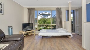 A modern hotel room with a large bed, a TV, a leather sofa, and a view of a house through the window. - The Lighthouse (Port Willunga)