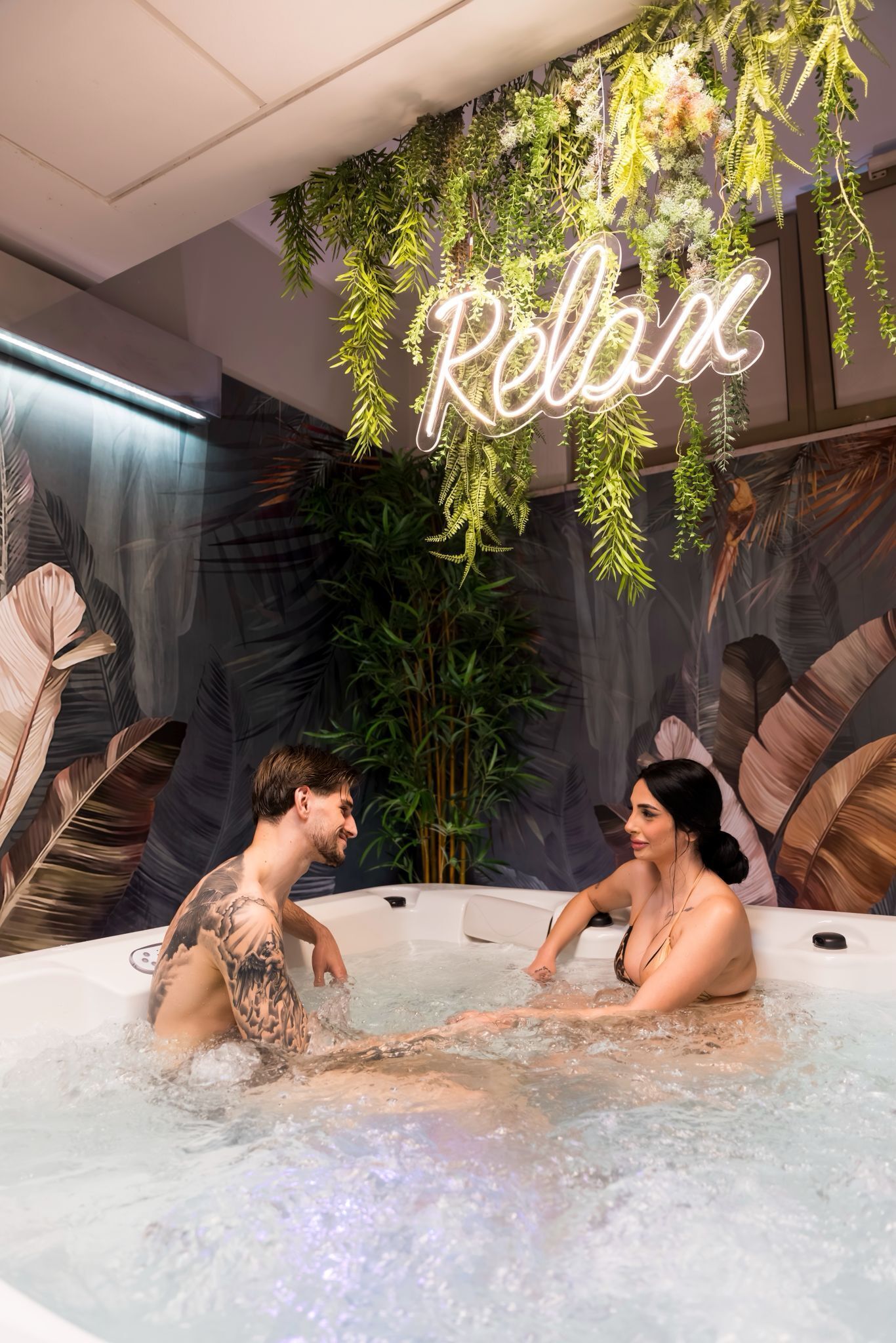 A couple in a hot tub with a neon sign reading 'Relax' and tropical foliage decorations.