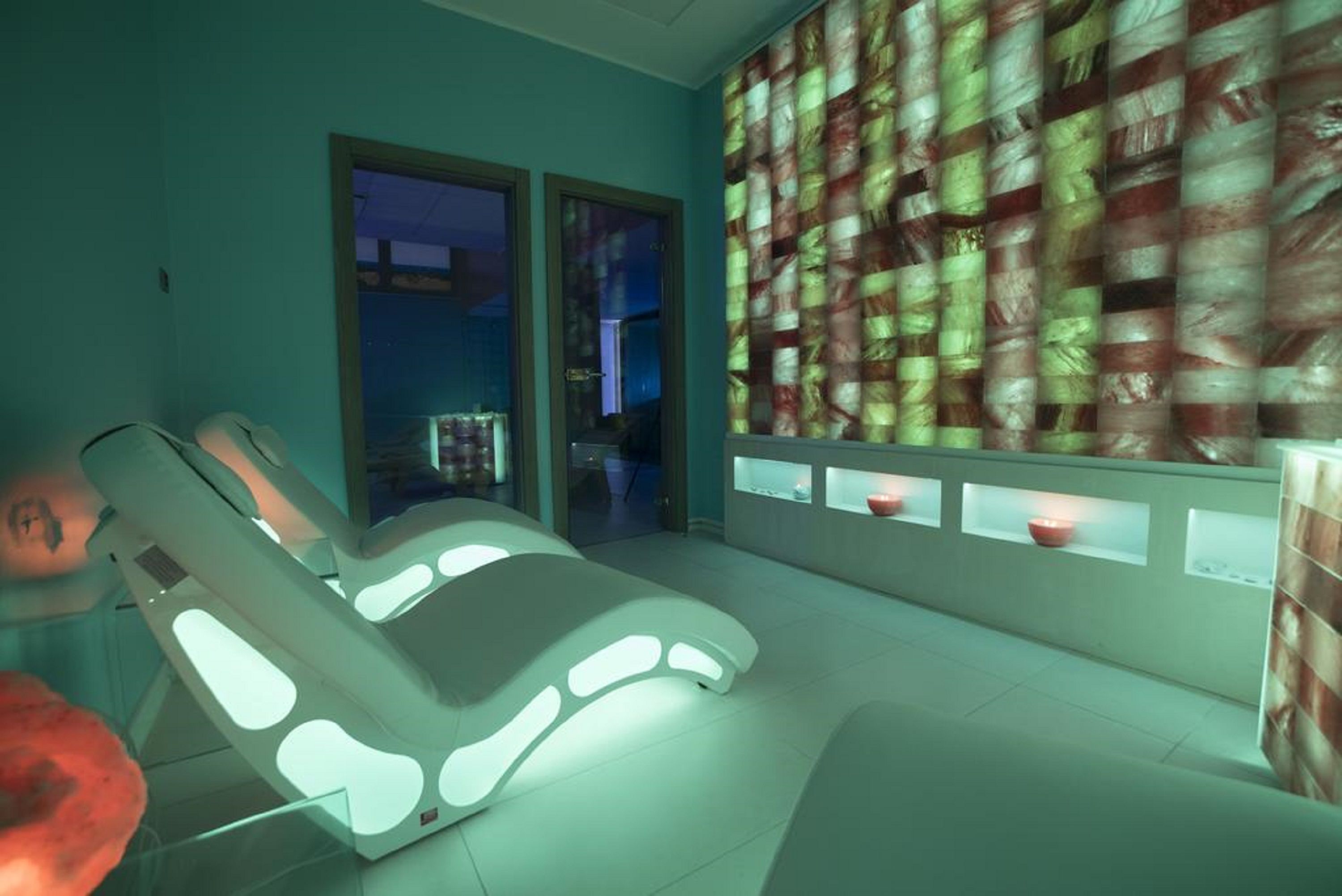 A modern spa room with a massage chair, a glass door leading to another room, and a patterned wall.