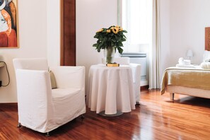 A hotel room with a white armchair, a small round table with a vase of flowers, a bed with a beige blanket, and a painting on the wall. - Barone Liberty & Luxury SPA-Boutique Hotel (Gallipoli)