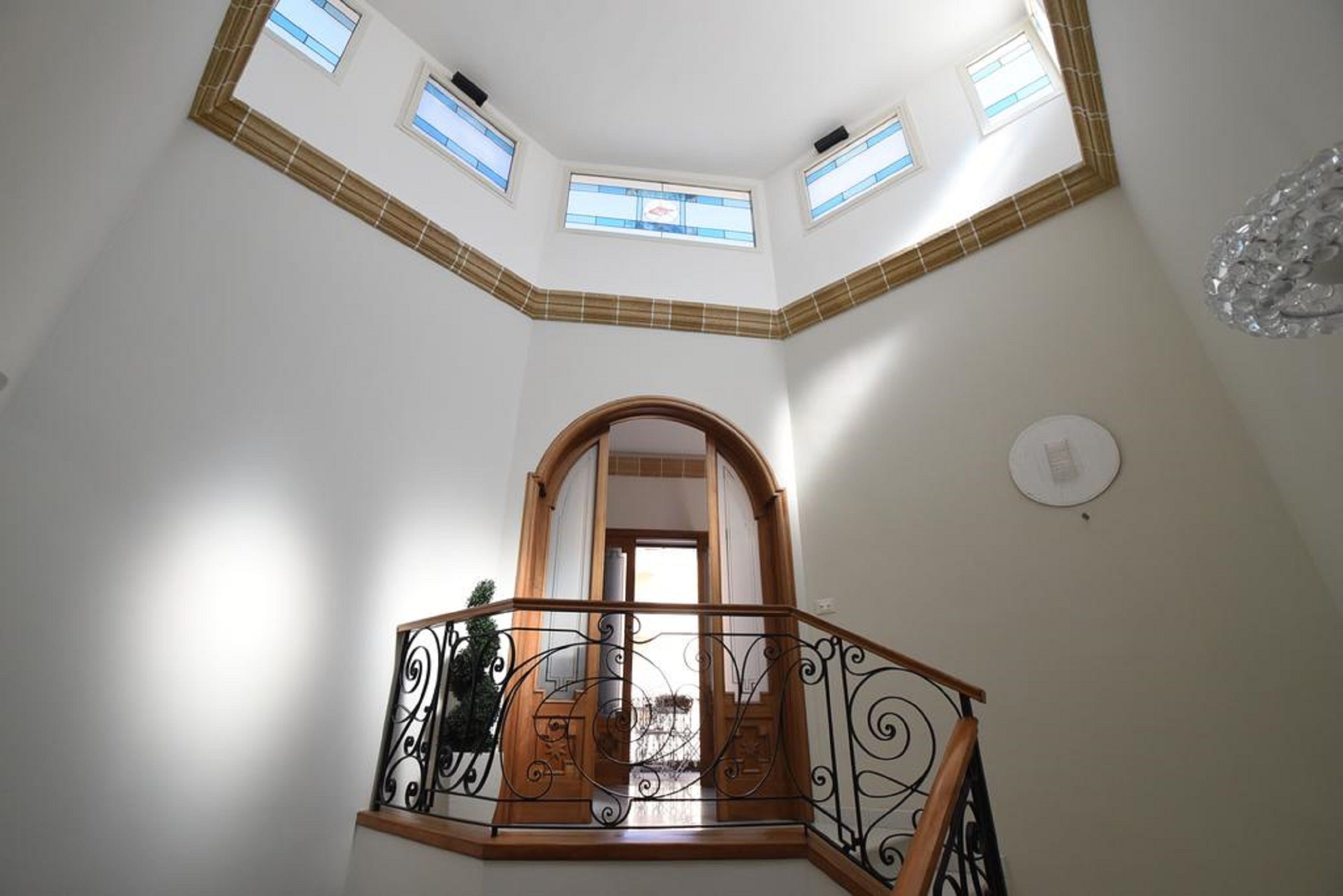 Tilava sisätila, jossa on korkea katto, koristeellinen mustilla kaiteilla varustettu puinen portaikko ja näkymä suuresta, kaarevan muotoisesta oviaukosta.