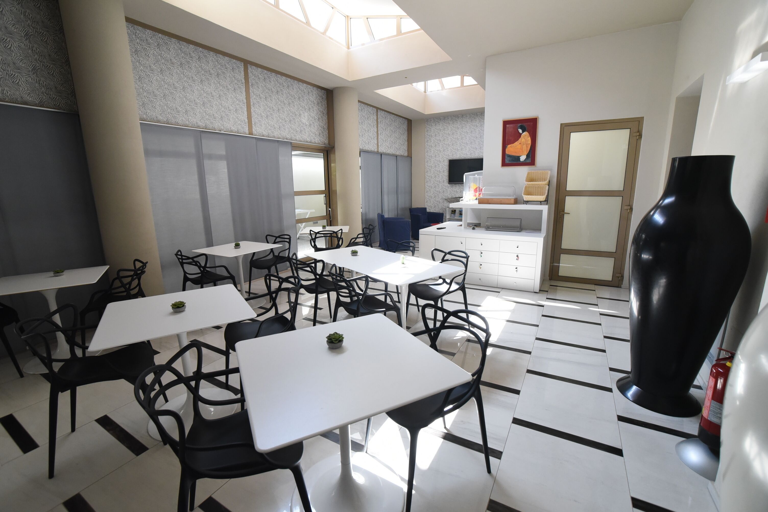 A modern dining area with white tables and black chairs, a kitchen area with a TV and a painting, and a large black vase on the right.