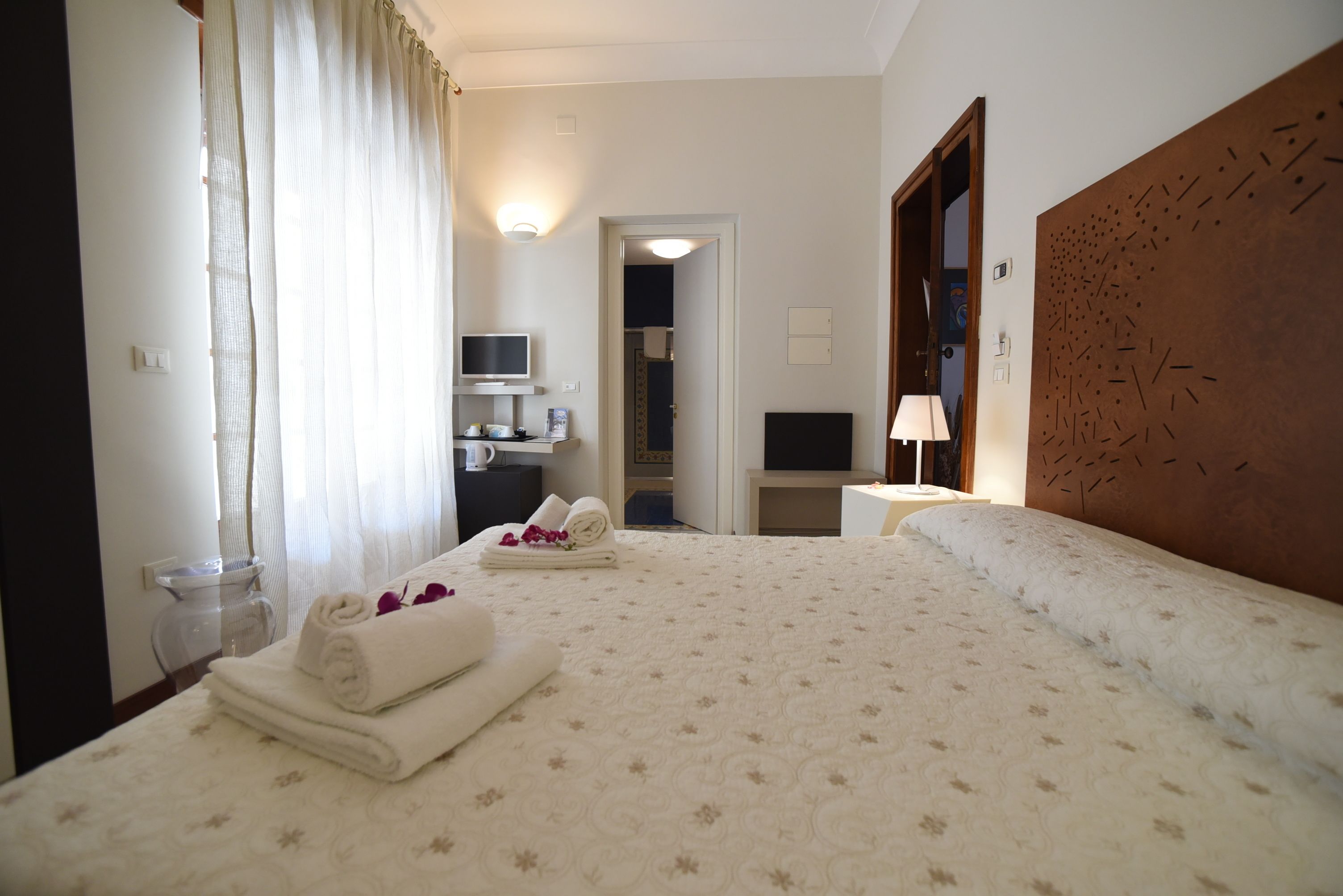 A hotel room with a large bed, a desk with a computer, a TV, and a bathroom visible through an open door.