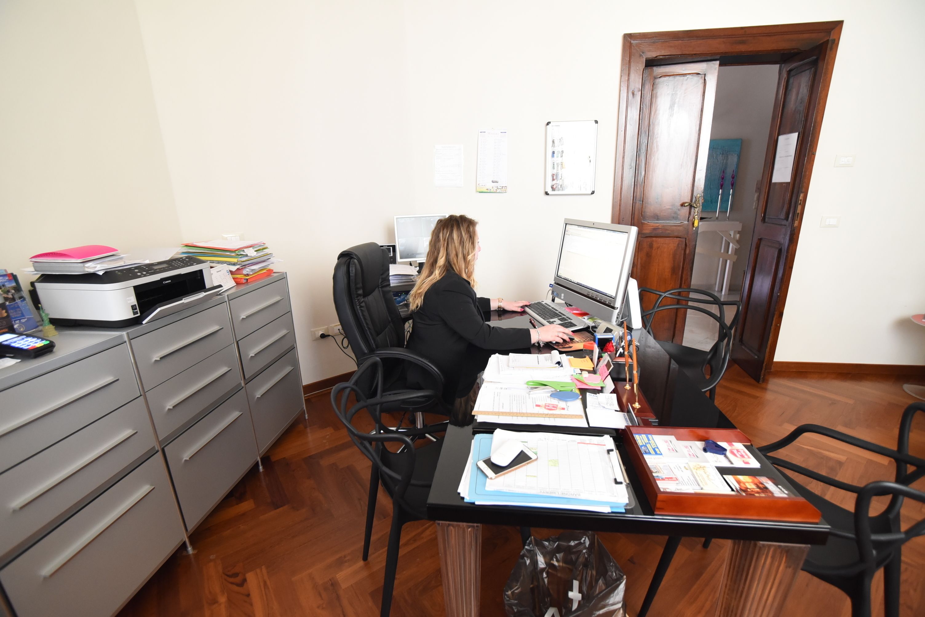 A room with a desk, computer, and various office supplies. There is a chair, a filing cabinet, and a door in the background.