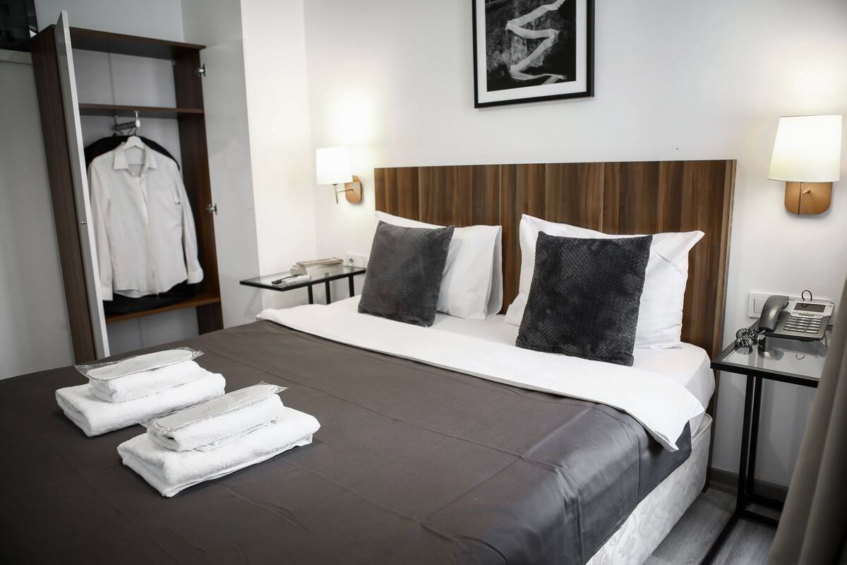 A hotel room with a bed, two bedside lamps, a nightstand, a phone, a framed picture on the wall, and a wardrobe with a hanging garment.