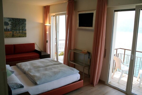 A hotel room with a bed, a red sofa, a TV, and a balcony with a chair. - Hotel Villa Carmen (Malcesine)