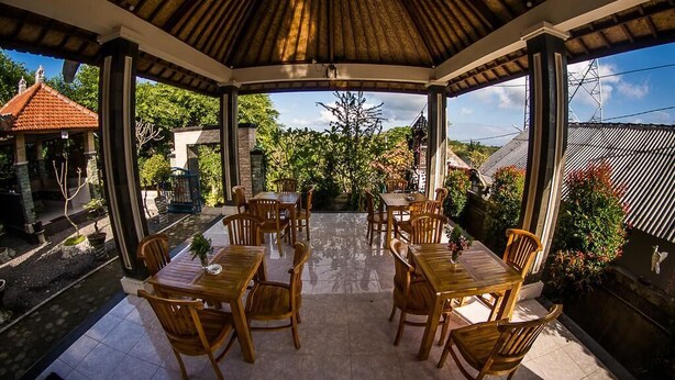 Un espacio de comedor al aire libre con techo, con mesas y sillas de madera, rodeado de follaje y edificios.