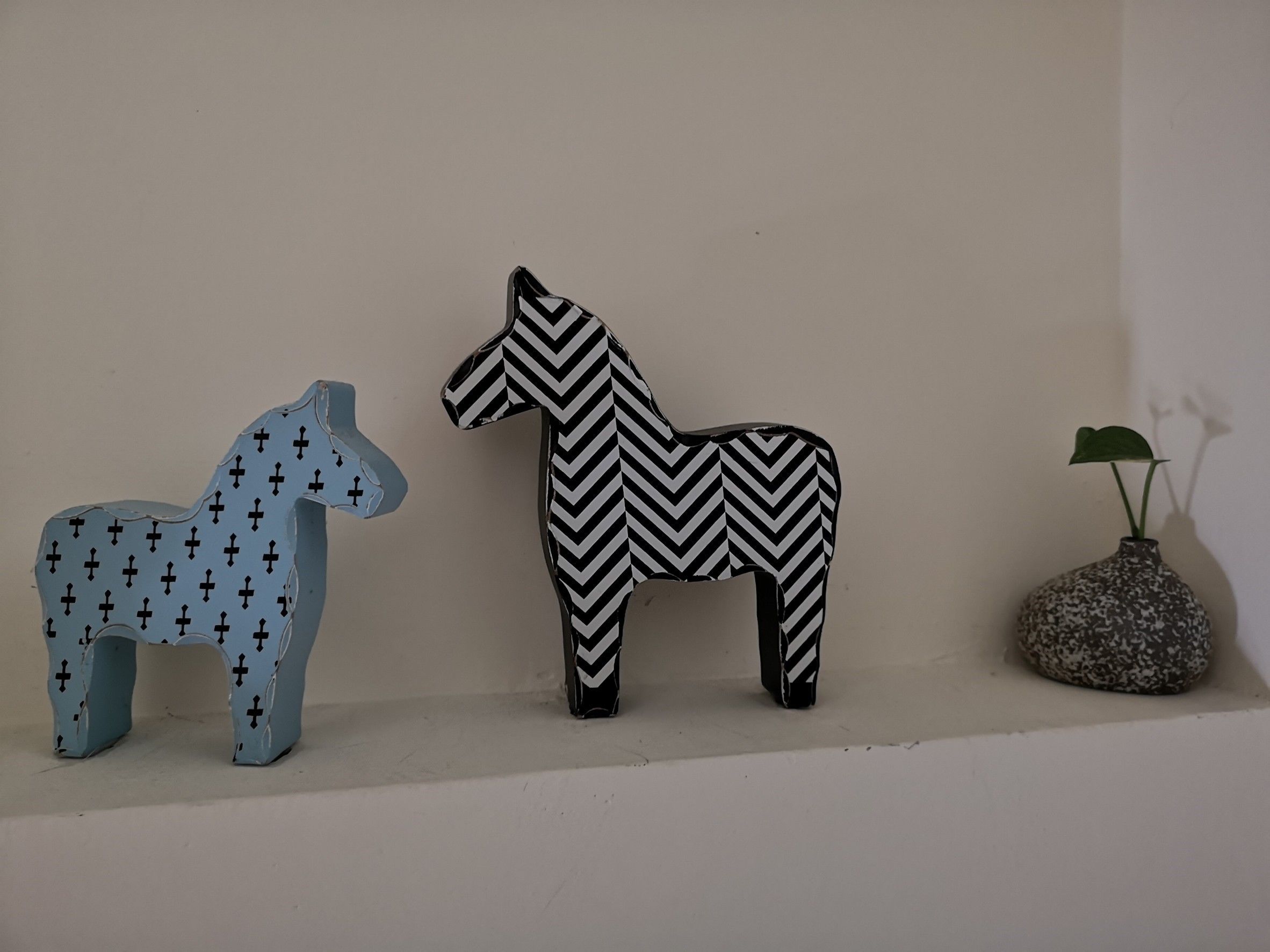 Two wooden horse-shaped sculptures on a shelf, one with a cross pattern and the other with a chevron pattern, next to a small plant in a rock planter.