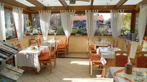 Un restaurante con terraza, cubierto por una estructura de madera, y mesas con manteles blancos y cojines anaranjados.