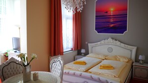 Una habitación de hotel con cama, candelabro, pintura de un atardecer, mesa de comedor y una silla.