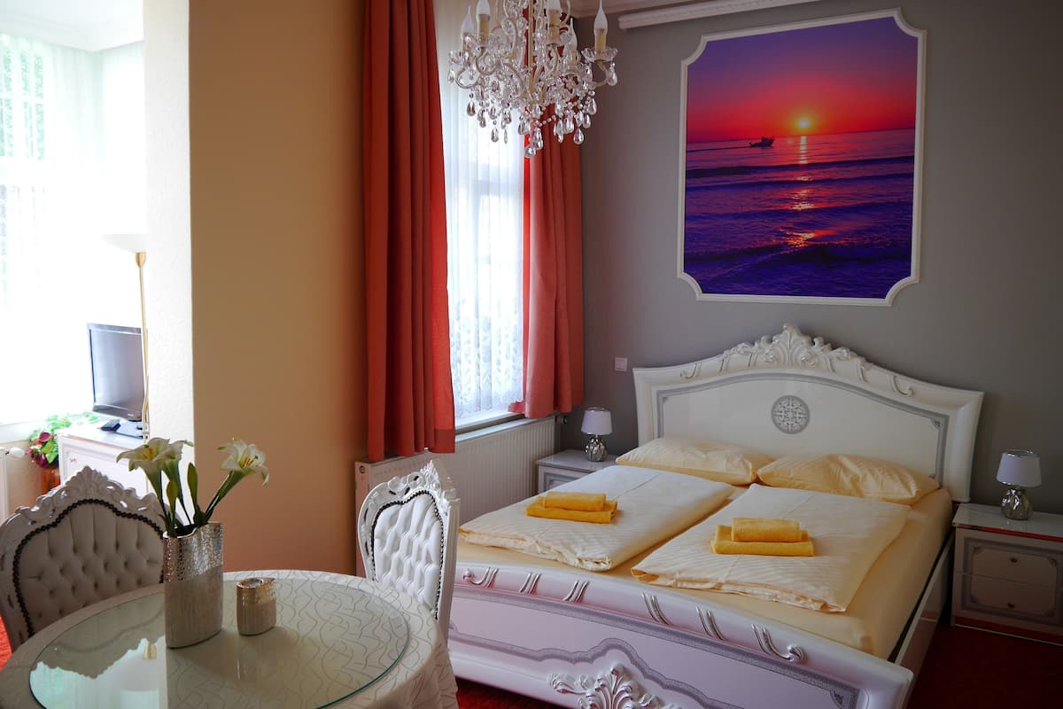 A hotel room with a bed, a chandelier, a painting of a sunset, a dining table, and a chair.