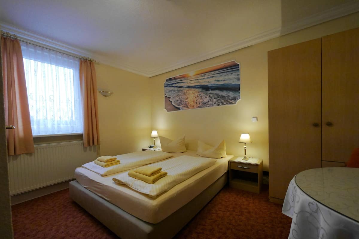 A hotel room with a bed, two bedside tables with lamps, a window with curtains, a painting on the wall, and a wardrobe.